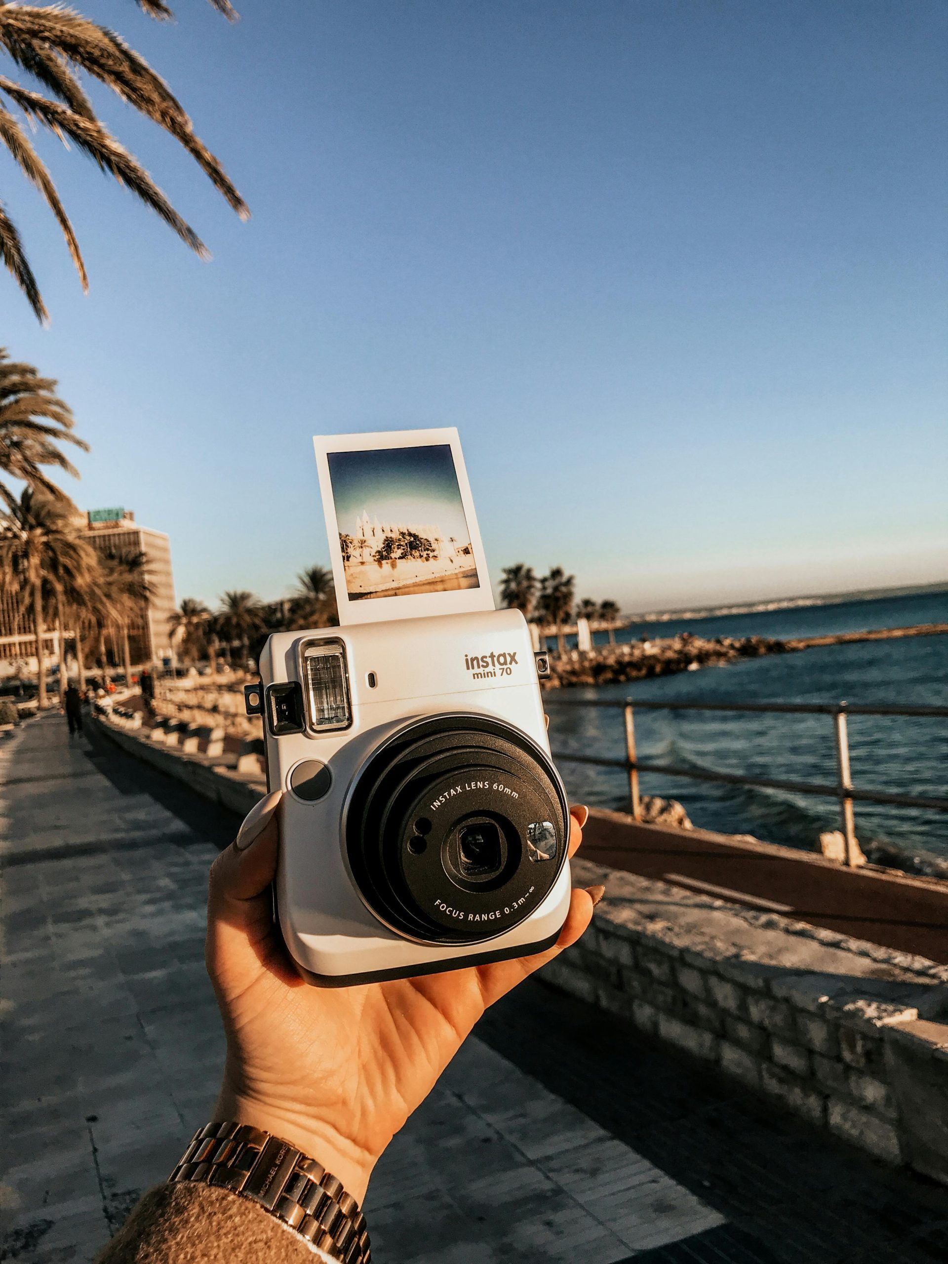 Close-up of a hand holding an Instax camera with seaside view and palm trees.