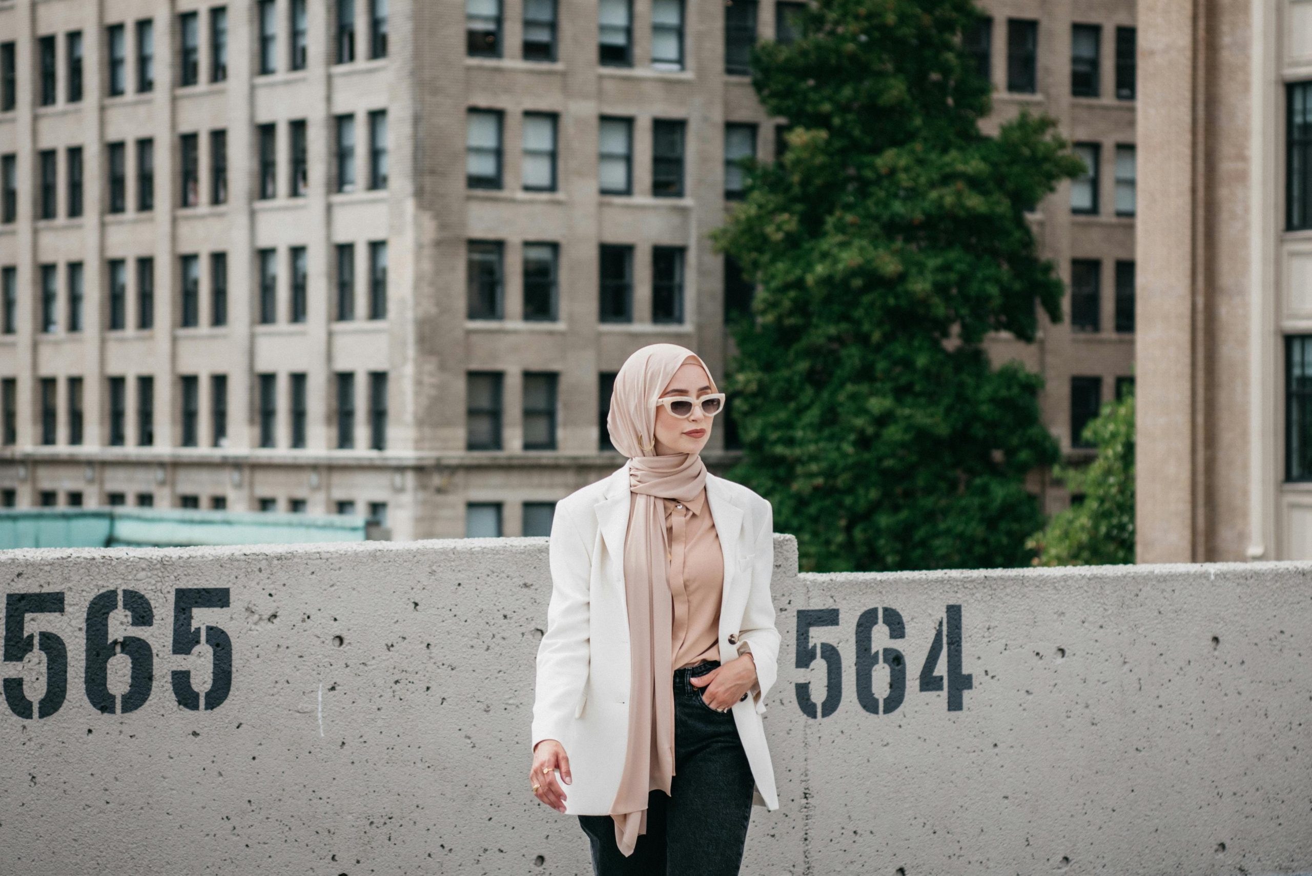 Stylish woman wearing sunglasses and hijab poses with city architecture backdrop.